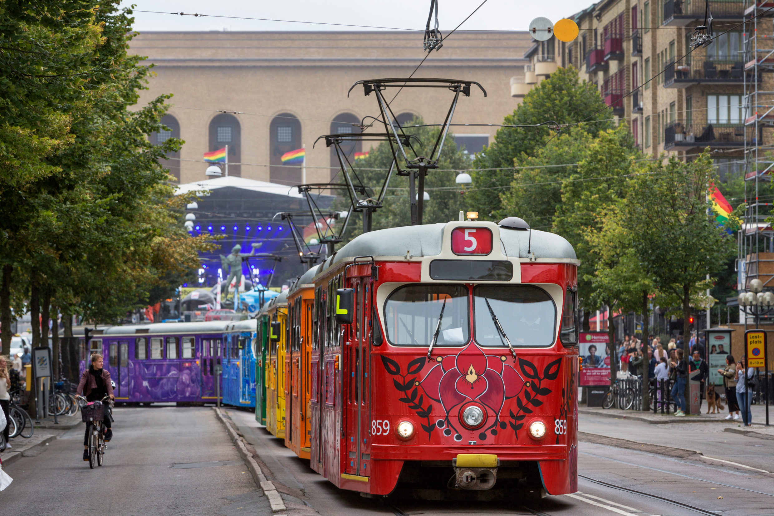 A tram going through a city is painted in colourful flowers, each car with one colour of the rainbow.